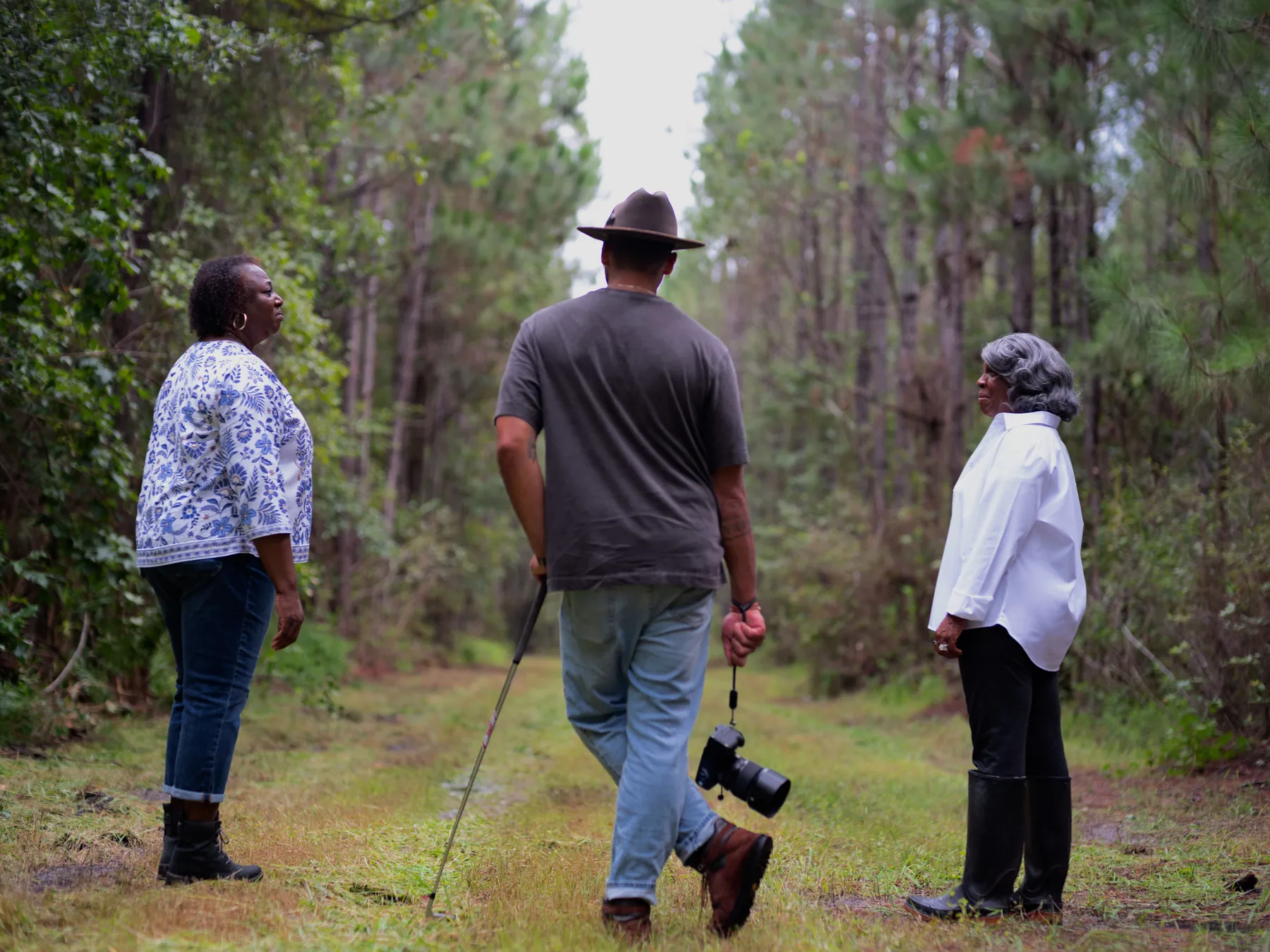 Marche Jenkins and her mother stand facing each other on a grass track through the pines as a photographer with a camera and monopod walks the line between them